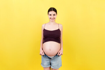 Happy pregnant woman in unzipped jeans showing her naked belly at colorful background with copy space. Baby expecting concept