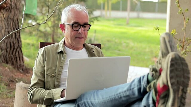 Gray-haired middle aged man using laptop outdoors. Man put feet on table and relax during a break