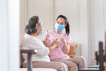 Young Asian daughter wearing surgical face mask to her mother sitting in living room at home. Online learning on laptop computer. Protect against coronavirus