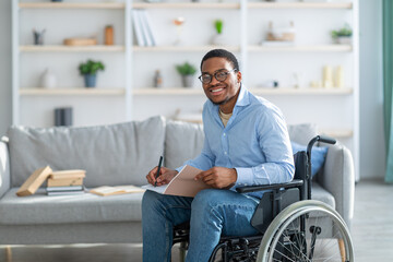 Happy disabled black guy in wheelchair taking notes, looking at camera and smiling at home, copy...