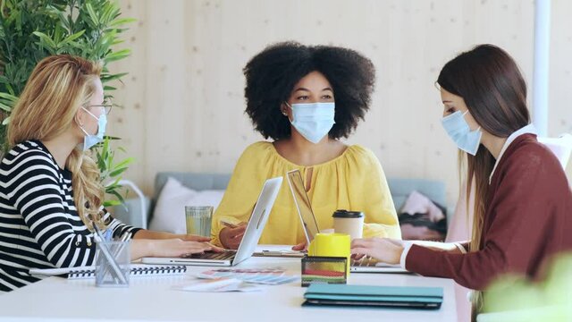 Video of multiethnic business women wearing a surgical mask working with laptops while talking of they new projects together in coworking place.