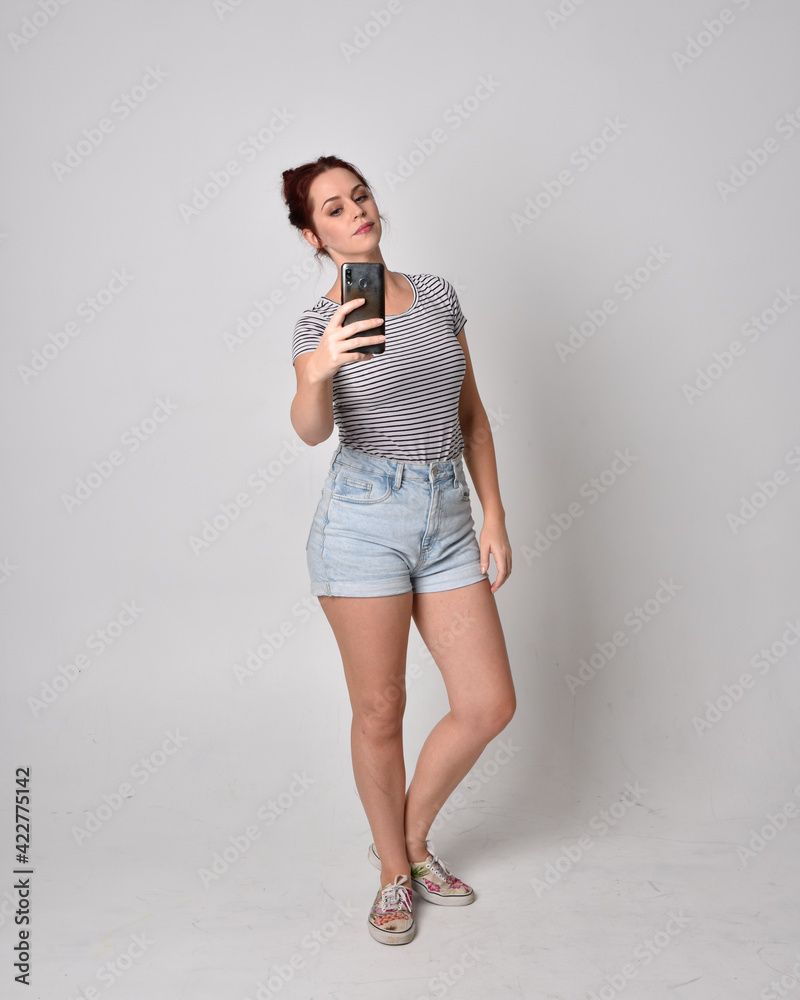 Sticker full length  portrait of girl wearing shirt and denim shorts. Standing pose holding a phone isolated on grey studio background.