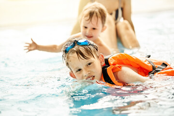 Boy with googles and safety vest learns to swim near brother and mom in large pool with clear water...
