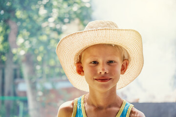 Cheerful little boy wearing large straw hat and blue sleeveless shirt posing for camera in sunny summer city park close view