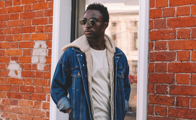 Fototapeta na wymiar Portrait of stylish young african man model wearing denim jacket posing on a city street over brick wall background