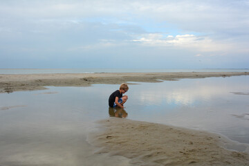 Boy at low tide on the beach, looks for small fish in the water in the evening