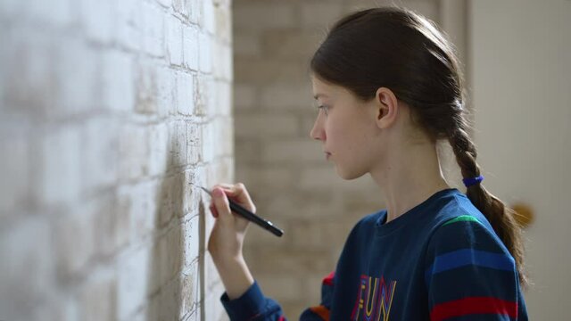 Girl with braid wearing fun sweatshirt solving mathematic examples on wallpaper with brick wall print. Side view schoolchild doing homework on unusual surface. Concept of education