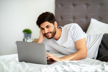 Cheerful arab man browsing internet on laptop while lying on bed at home, using computer in morning