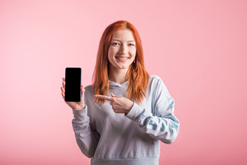 Naklejka premium Redhead teenager girl points with finger at smartphone blank screen in studio on pink background