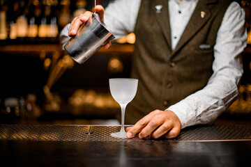 view of glass on bar over which the bartender's hand holds steel mixing cup