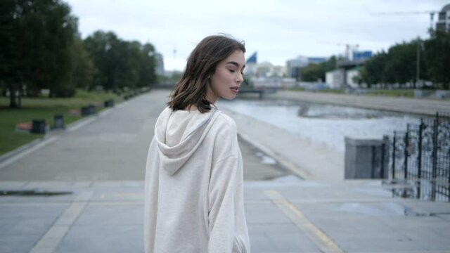 Attractive young brunette girl in a white hoodie walks along the city's waterfront and turns on camera. High quality FullHD footage