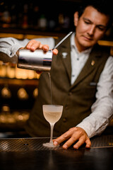 bartender pours cocktail from steel mixing cup with strainer into glass