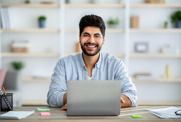 Positive arab male employee smiling at camera while working on laptop at home office