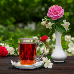 Glass cup with tea among flowering bushes of roses and jasmine. Outdoor, picnic, brunch. Green leaves background in blur.