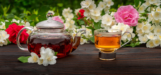 A steamed teapot and an elegant glass cup among flowering rose and jasmine bushes. Outdoor, picnic, brunch. Green leaves background in blur.