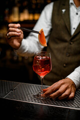 bartender gently holding tweezers with citrus slice over wine glass with iced cocktail