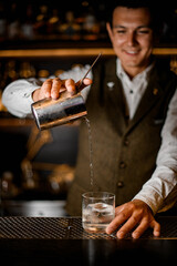 professional bartender pours drink from steel mixing cup with strainer into old-fashioned glass with ice