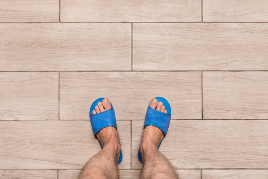 Men's Bare Feet In Blue Home Slippers Stand On A Light Laminate Floor Tile Background, View From Above