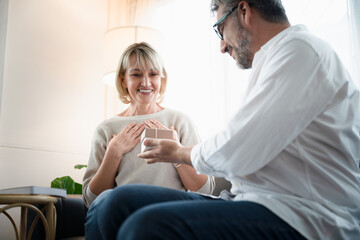 Obraz premium Elderly old man give a present to old woman in the warm light atmosphere, Romantic elderly husband surprise his wife with a present for anniversary. Happy senior people lifestyle at home.