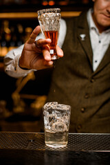 man bartender holds inverted crystal bottle over transparent glass with ice cubes