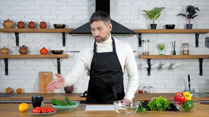Man professional chef in apron standing near table in home kitchen tells shows ingredients for cooking salad, teaches housewife student records video online remote webinar master class culinary course