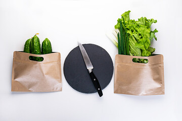 Fresh green vegetables, salad, green onions and cucumbers in reusable paper bags and a board with a knife on a white background, top view. Healthy food, food for vegetarians. Zero west concept.