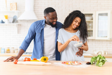 Obraz na płótnie Canvas Happy african american couple cooking dinner together, trying new recipe where the main ingredient is love, woman adding grated cheese on dough for making delicious Italian pizza, husband supports her