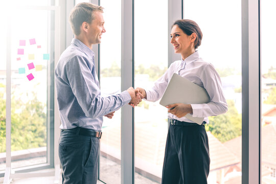 Handshake successful business together, portrait of young caucasian businessman and businesswoman happy smiling standing near window in office. Partnership finance teamwork contract business concept.

