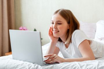 Young woman smile and work using laptop while lying in bed in morning at home. Happy girl in pajamas studying online shopping or planning her day use computer laptop in morning time in bedroom.