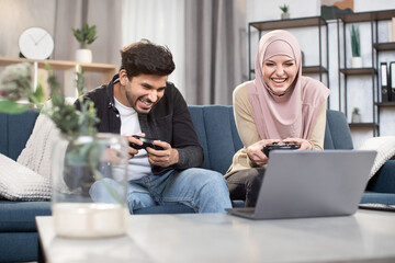 Laughing 30-aged funny Muslim couple having fun together while playing video games at home using gamepads. Family leisure, playing video games.