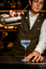 Fototapeta na wymiar man bartender holds mixing cup with strainer and pours blue cocktail into glass on bar