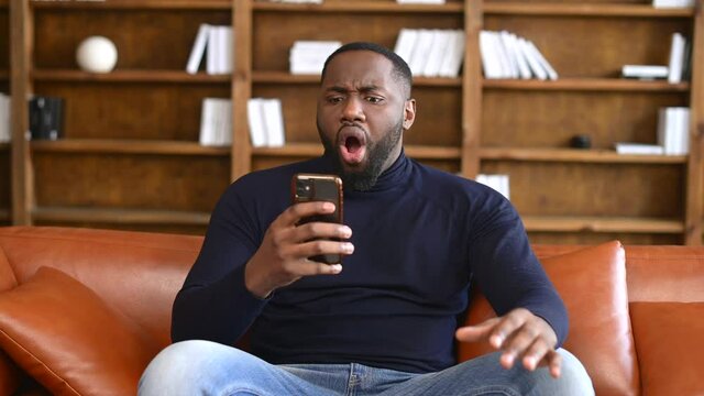 Shocked African-American man holding a smartphone, feel astonishment with a bad news, fired from work, irritated guy looks at phone screen and does not understand what happened