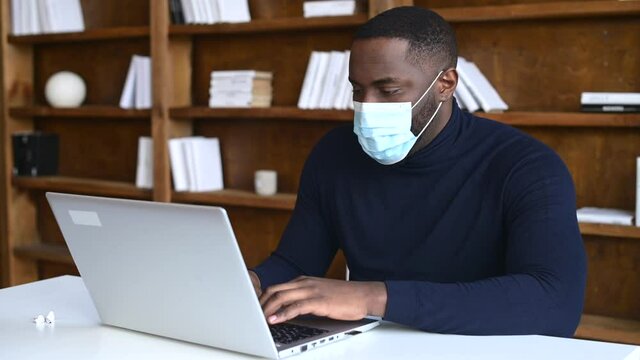 Focused African-American wearing medical mask working with a laptop in contemporary office space with bookshelves on the background, social distancing and protective measures for office employees