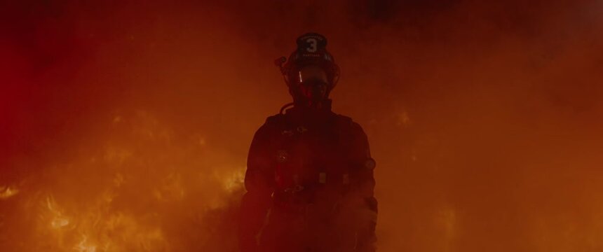 Dramatic shot of American firefighter in full gear walking through smoke away from huge fire. Shot with 2x anamorphic lens 