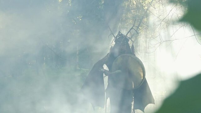 Shaman walks in the mixed forest and plays the drum. Lot of smoke. Sun rays.Slow motion.