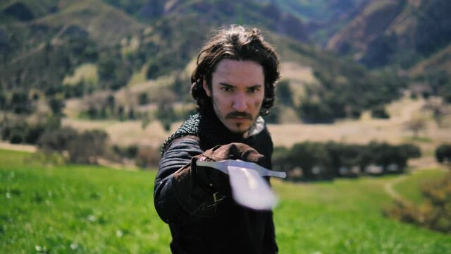 Man in medieval outfit draws sword from scabbard and points it at the camera. camera tracks backward, 4k, 60fps slow motion