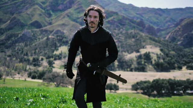 Man in medieval outfit walks forward with mountains in the background. 60fps, slow motion, camera tracking back
