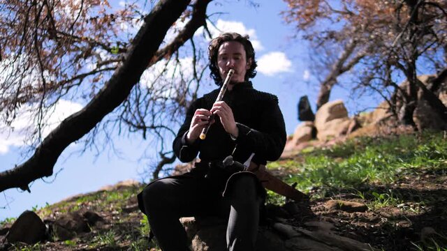 Man in medieval outfit sitting on a rock playing the flute from the front.  He begins to rock back and forth. 4k, tracking camera, 60fps, slow motion
