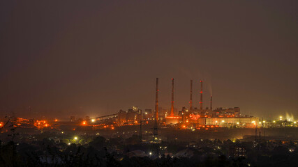 Night urban landscape with an industrial complex with its metal structures and chimneys with reflection of lamps with yellow lights of the city.