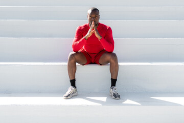 African sportsman with red sportswear sitting on white stairways in a stadium.