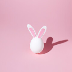 White egg with fluffy bunny ears and summer shadow on pastel pink background. Creative Easter fun...