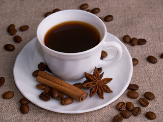 A white cup of espresso stands on a burlap table with scattered coffee beans, a cinnamon stick and stars of anise.