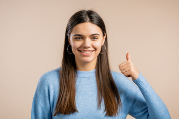 Woman showing like thumb up sign over beige background. Positive young girl smiles to camera. Winner. Success. Body language.