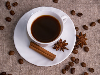 A white cup of espresso stands on a burlap table with scattered coffee beans, a cinnamon stick and stars of anise.