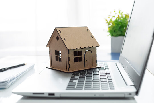 Small wooden house model standing on a laptop keyboard on a bright office desk. Online real estate searching, home insurance, and property investment concept.