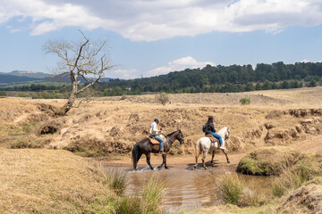 Dos jóvenes con sus caballos están cruzando un arroyo. © jesuschurion57