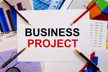 The words BUSINESS PROJECT is written on a white background near colored graphs, pens and pencils. Business concept