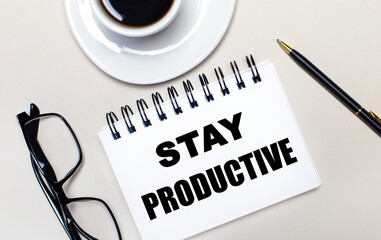 Glasses, a white cup of coffee, a white notebook with the words STAY PRODUCTIVE and a ballpoint pen lie on a light background. Flat lay. View from above.