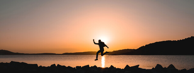 Horizontal banner silhouette of traveler on seashore jumping at the beach during sunset.