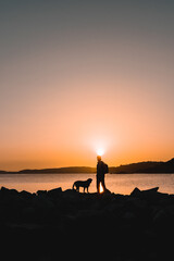 Silhouette of hiker man standing on the rocks in a coastline on the beach with his dog. Background with beautiful sunset.
